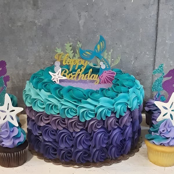 Mermaid-themed birthday cake with teal and purple frosting rosettes, starfish, and cupcakes.