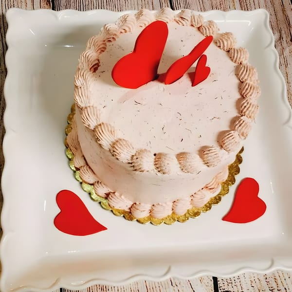A pink frosted cake with red heart decorations sits on a white platter.
