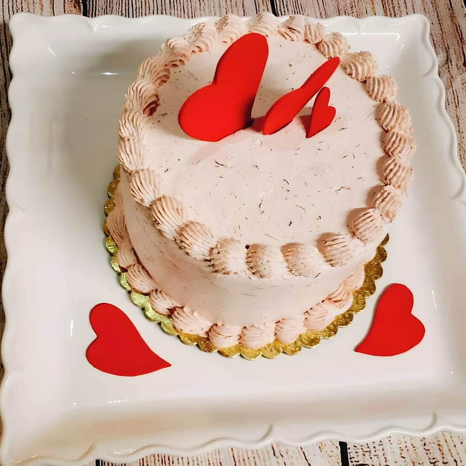 A pink frosted cake with red heart decorations sits on a white platter.