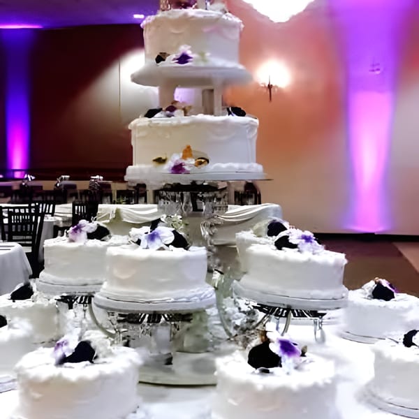 A multi-tiered white wedding cake and several smaller cakes adorned with purple flowers in a banquet hall.