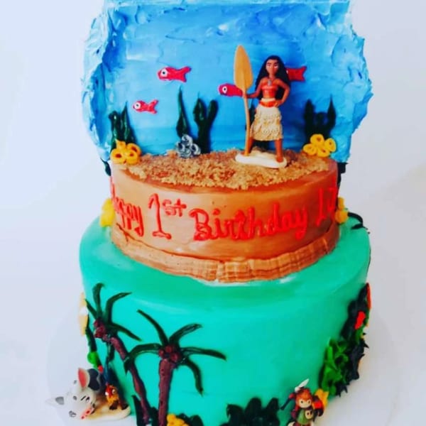 Two-tier Moana-themed birthday cake with a Moana figurine, palm trees, and "Happy 1st Birthday" text.