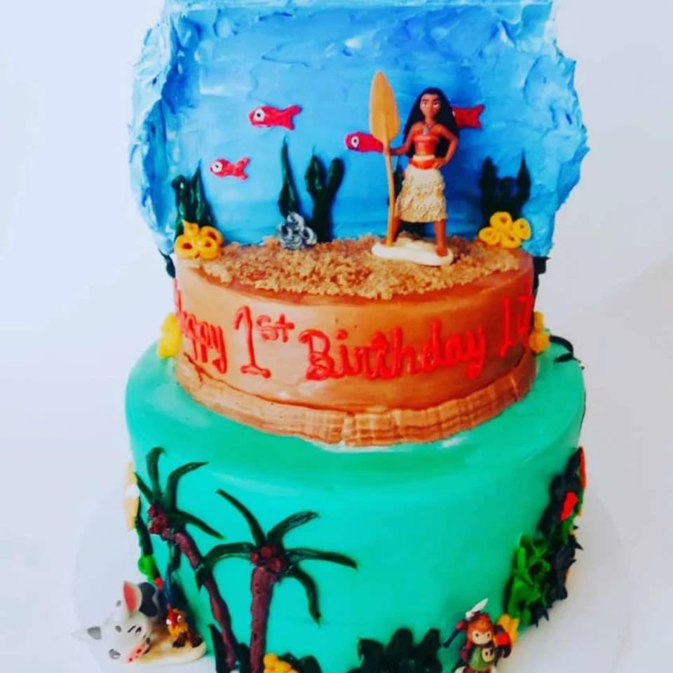 Two-tier Moana-themed birthday cake with a Moana figurine, palm trees, and "Happy 1st Birthday" text.