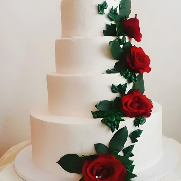 A four-tiered white wedding cake adorned with red roses and green leaves cascading down the side.