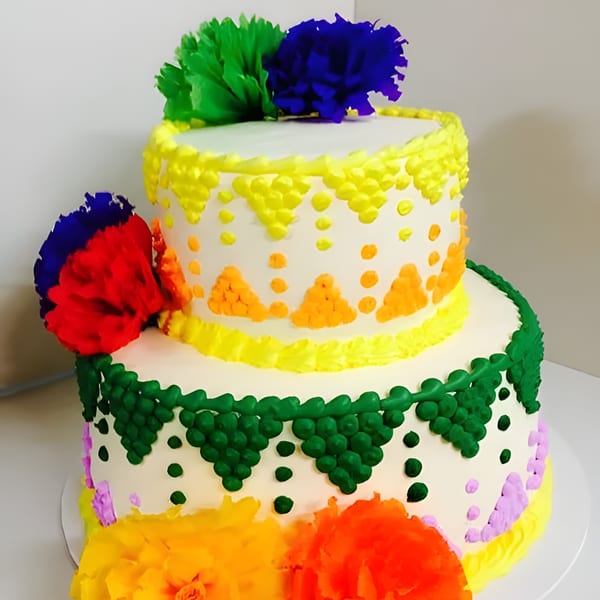 Two-tiered white cake with colorful Mexican-inspired frosting designs and paper flower decorations.