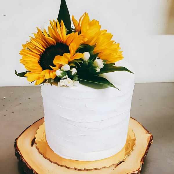White frosted cake topped with vibrant sunflowers and small white flowers on a wooden slab.
