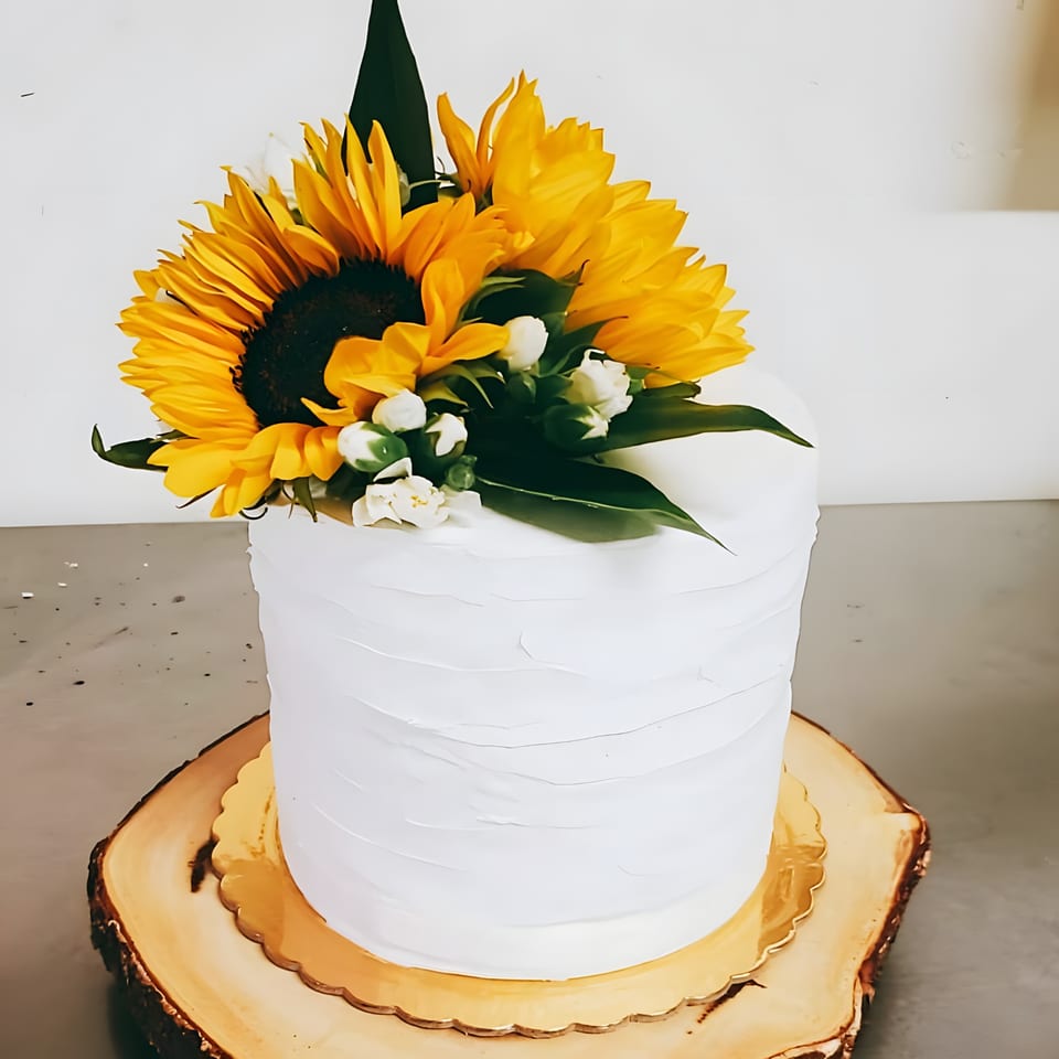 White frosted cake topped with vibrant sunflowers and small white flowers on a wooden slab.