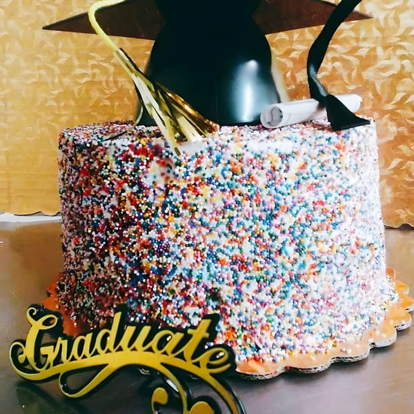 A colorful sprinkle-covered graduation cake with a black cap, diploma, and "Graduate" sign.